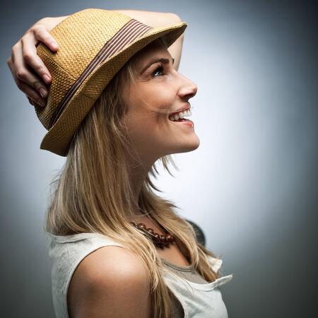 Close up Side View of Happy Blond Woman in Trendy Attire, Wearing Sleeveless Top and Brown Necklace While Holding her Hat on Head. Captured in Studio with Gradient Gray Background.の写真素材