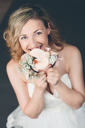 Tender shy young bride hiding in her bouquet of flowers looking up at the camera with a radiant smile of happiness, studio portrait on greyの写真素材