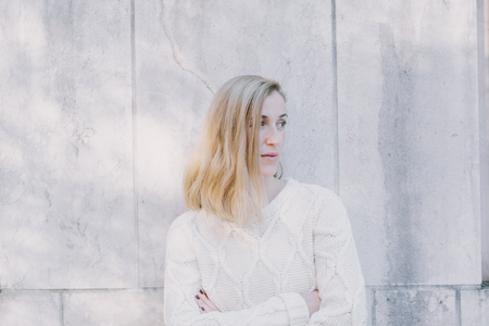 Serious attractive blond woman standing with her arms crossed staring to the right side of the frame watching something, white stone wall background with copyspaceの写真素材