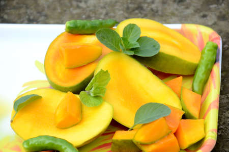closeup the sliced ripe green yellow mango fruit with green mint leaves and chilly in the tray over out of focus grey brown background.の写真素材