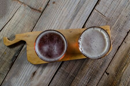 Two glasses of dark amber beer against a wooden rustic cuting board on textured background, top...