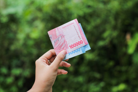 A woman's hand holds a rupiah banknote against a nature background. 150000 Rupiah.