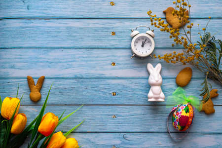 Easter card, party supplies, mimosa flowers and yellow tulips on a light blue wooden background. Handmade cookies in the shape of a bunny, chicken, colored bright eggs. Alarm clock reminder of the eve of the Easter holiday. Place for text.の写真素材