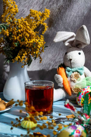 Greeting Easter card. A hot drink in a glass, a cheerful bunny with a carrot, festive colorful eggs,...