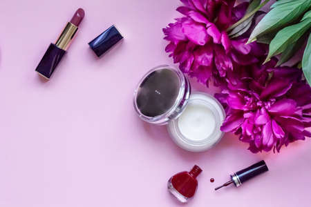 Close-up flat lay on pink background with place for text beauty products, lipstick, face cream and bright nail polish, large red peony flowers.の写真素材