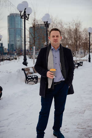 A young businessman in a coat and suit stands on the alley of the city with coffee, looks at the...