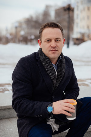 Close-up portrait of a young handsome man sitting on concrete steps with coffee in a city park with...