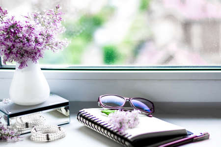 Cozy home still life: spring lilac flowers in a white vase on the windowsill, notebook and glasses. vintage style.の写真素材