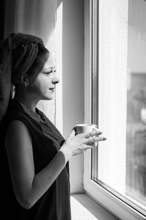 A young beautiful girl after a shower in a towel on her head, stands at home looks out the window...