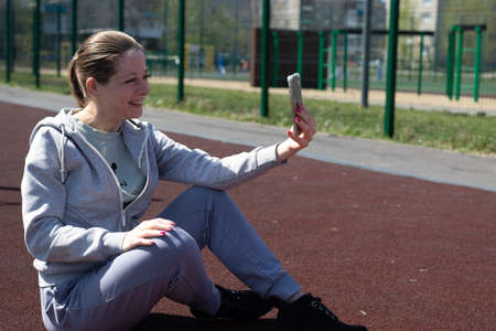 A young blonde girl in sportswear is resting after doing sports, running, sitting at the stadium, talking on a mobile phone, social networks.の写真素材