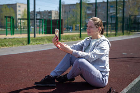A young blonde girl in sportswear is resting after doing sports running, sitting at the stadium, talking on a mobile phone video chat.の写真素材