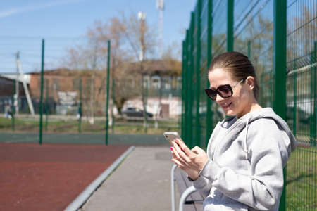 A young blonde woman in sportswear is resting after playing sports, running, sitting on a bench in the stadium, communicating in social networks.の写真素材