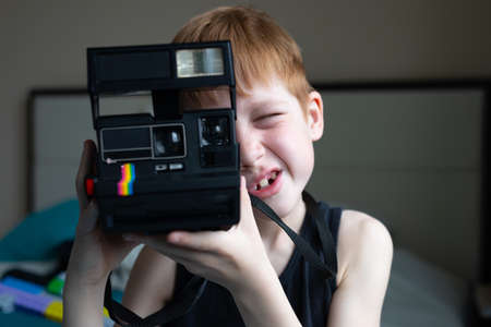 Child red-haired boy holds an old retro camera in his hands and makes a photo with a smile, children's hobby photography, life style portrait at home.の写真素材