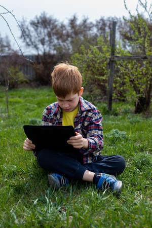 Happy child redhead boy playing on tablet or watching cartoons sitting on green grass in the backyard in the village.の写真素材