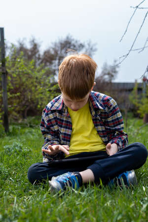 Happy child redhead boy playing on tablet or watching cartoons on green grass in the backyard in the village.の写真素材