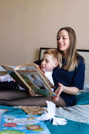 Family holiday mother with a red-haired child boy son watching a folded album from kindergarten at home in the bedroom on the bed.の写真素材