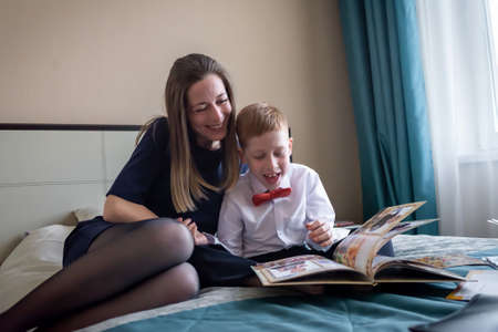 Family holiday cheerful young mother with a red-haired child boy son watching a folded album from kindergarten at home on the bed.の写真素材
