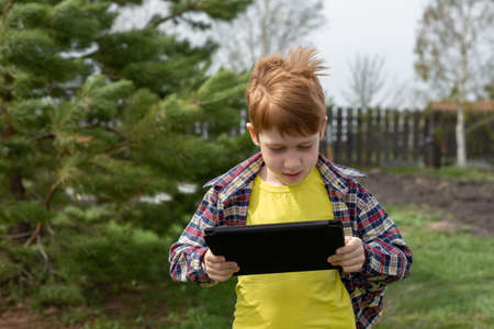 Happy redhead boy playing tablet or watching cartoons, walking in the backyard in the village, summer vacation.の写真素材