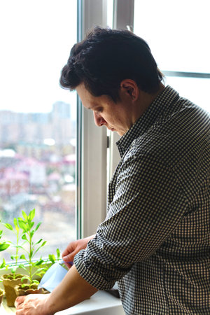 Home plant care concept. Young man watering seedlings on the windowsill, homework routine.