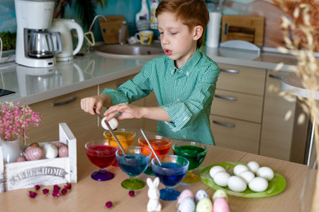 A beautiful sweet and funny red-haired boy paints eggs in the kitchen for Easter. Colored eggs, the child paints at home, ortrait.の写真素材