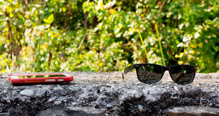 Mobile phone and sunglasses against a stone and trees background. Perfect for showcasing tech during a hike or in outdoorsの写真素材