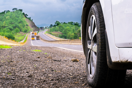 Low angle shot of a car wheel against a hill road bordered by trees with trucks passing itの写真素材