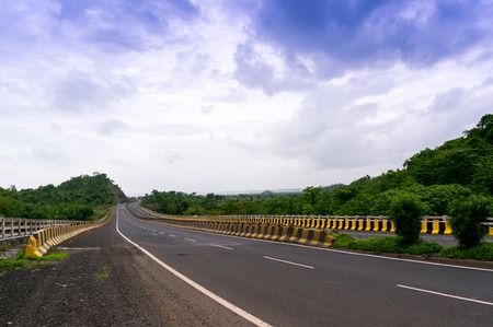 Long stretch of empty road bordered with tree filled hills and under beautiful monsoon skies. Showing a great way to explore india via the well developed road networkの写真素材