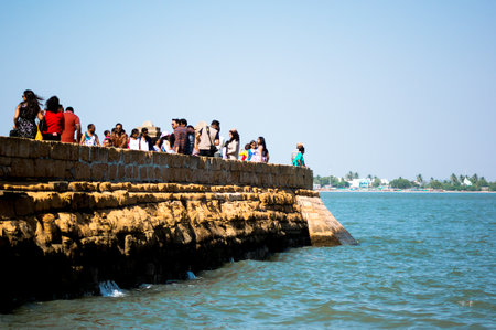 Diu, Gujarat, India - Circa 2018 : People, men, women, children walking on a stone jetty at Diu fort with Pani Kotha prison in the distance. Shows the blue water, waves, ocean around this famous monument, landmark and tourist spot with the stone walls of のeditorial素材