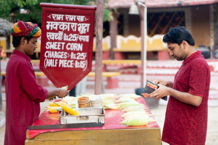 Traditionally dressed rajasthani vendor serving corn on cob to a young man at a festival in Indiaのeditorial素材