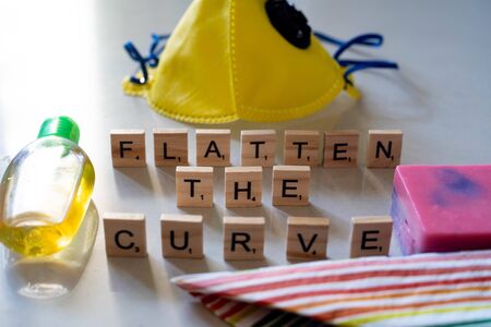 Flatlay with wooden blocks spelling flatten the curve, a yellow face mask, sanitizer and soap for...