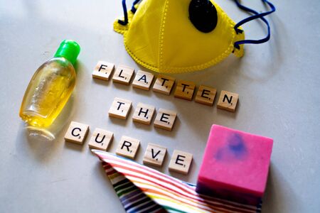 Flatlay with wooden blocks spelling flatten the curve, a yellow face mask, sanitizer and soap for coronavirus safetyの写真素材