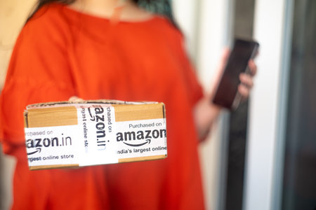 Young indian girl holding an amazon delivered box in one hand an an out of focus mobile phone in the other showing a delivery from this e-com vendorのeditorial素材