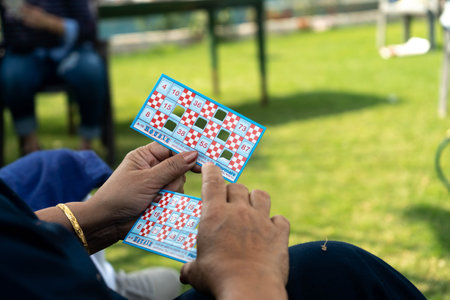 Woman holding a tambola housie lotto bingo ticket in one hand with some numbers folded while sitting in a gardenのeditorial素材