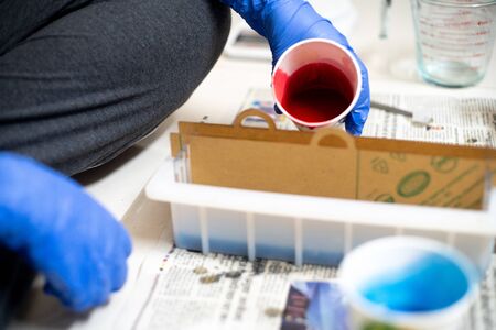 Blue gloved hand pouring red liquid from a paper cup into a mold with partitions for making a home...