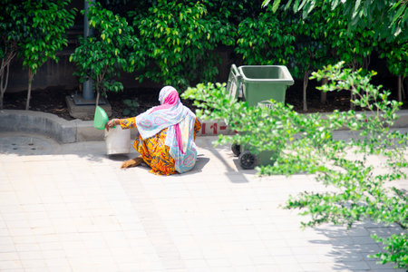 Old lady in saree and mask cleaning pavements sidewalks and streets with a wooden broom and putting...