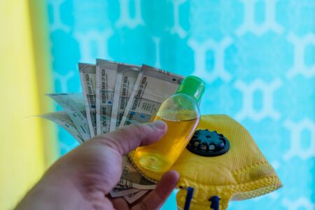 Hand holding out indian currency notes,yellow mask and sanitizer, against a colorful backgroundの写真素材