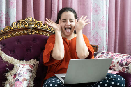 Young indian woman in a red dress sitting cross legged on an ornate sofa with elegant curtains working on a laptop working from home in the coronavirus pandemic lockdownの写真素材