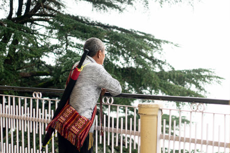 Buddhist monk in civilian clothes standing in a sweater with a bag and umbrella looking at the fog covered trees in Dharamshala Mcleodganjのeditorial素材
