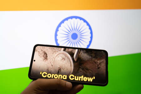 man holding a mobile phone with news on corona curfew and the india tri color flag in the background showing the self lockdown during the second stage of the pandemicの写真素材