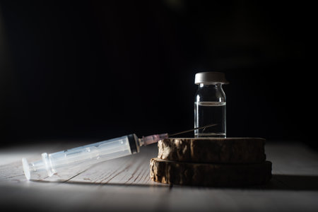 syringe pointing upwards with glass vial filled with liquid serum medicine on a wooden block shot against black concept of fake spurious counterfeit medicine being black marketed in the pandemicの写真素材