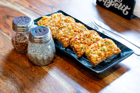 Garlic bread slices with butter mozzarella vegetable onion bell pepper on a black plate placed on a wooden table with salt pepper and oregano on the side in a delhi cafe in indiaの写真素材