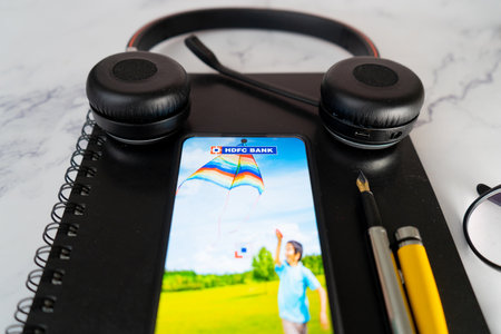 Smart phone with HDFC housing development finance corporation bank app with pen, headphone, notepad and specs showing saving and investments being done online in Indiaのeditorial素材