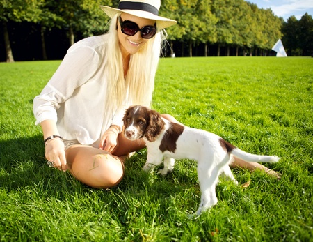 A stylish young blonde woman sits in lush grass playing with her dog in evening sunshine in a park. A stylish young blonde woman sits in lush grass playing with her dog in evening sunshine in a park.
