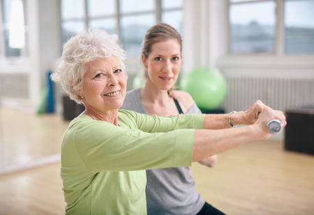 Senior woman exercising with fitness trainer at gym. Active senior woman lifting dumbbells with help from personal trainer. Senior woman exercising with fitness trainer at gym. Active senior woman lifting dumbbells with help from personal trainer.