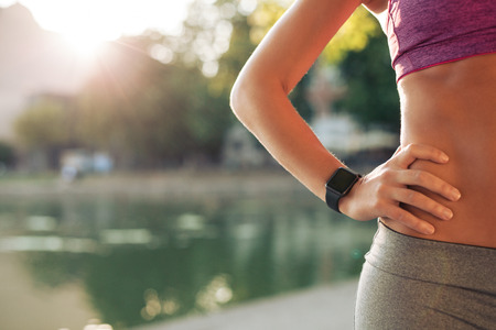 Sportswoman wearing smartwatch device. Cropped shot of fit woman in sports wear standing with her hand on hip outdoors, with sun flare. Sportswoman wearing smartwatch device. Cropped shot of fit woman in sports wear standing with her hand on hip outdoors, with sun flare.