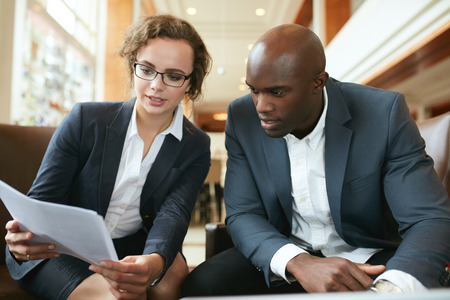 Two business partners sitting together and discussing contract documents. Business executives going through papers in lobby. Two business partners sitting together and discussing contract documents. Business executives going through papers in lobby.