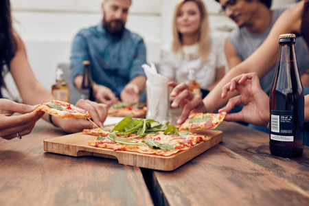 Close up shot of pizza on table, with group of young people sitting around and picking up a portion. Friends partying and eating pizza. Close up shot of pizza on table, with group of young people sitting around and picking up a portion. Friends partying and eating pizza.