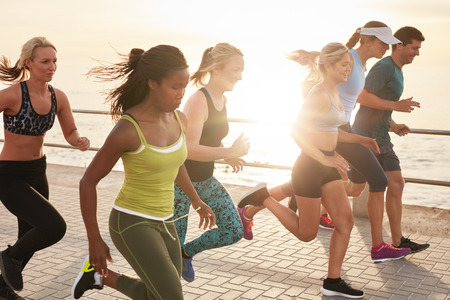 Portrait of healthy young men and women running race on seaside promenade. Group of young people sprinting outdoors at sunset. Portrait of healthy young men and women running race on seaside promenade. Group of young people sprinting outdoors at sunset.