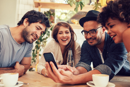 Group of young people sitting in a cafe and looking at the photos on smart phone. Young men and women meeting at cafe table and using cell phone Group of young people sitting in a cafe and looking at the photos on smart phone. Young men and women meeting at cafe table and using cell phone