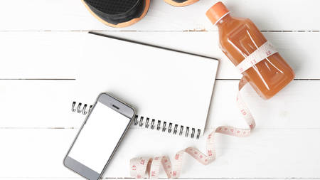 fitness equipment:running shoes,juice,measuring tape,notepad and phone on white wood background